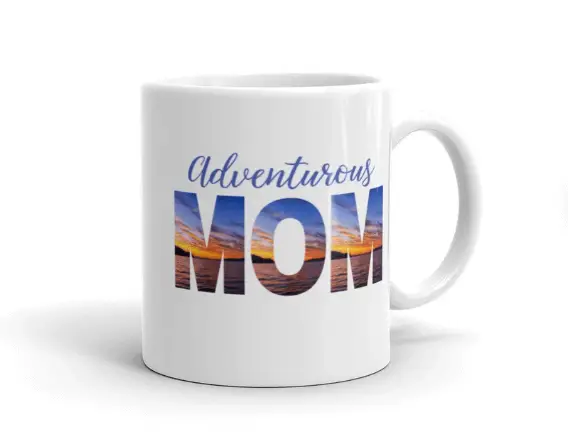 white mug, basic traditional style, in blue text "adventuous" in big block letters under it "mom" with a sunset printed inside the block letters